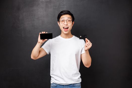 Bank, finance and payment concept. Enthusiastic young asian male student showing his new credit card and features of online banking application, standing amused over black background.