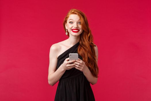 Portrait of excited and amused, redhead woman wearing black dress on her date or prom night, holding mobile phone, smiling beaming at camera, standing red background upbeat.