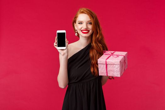 Portrait of elegant and stylish redhead woman showing mobile phone display and pink gift box, recommend store or delivery service to buy presents for your girl, smiling, red background.