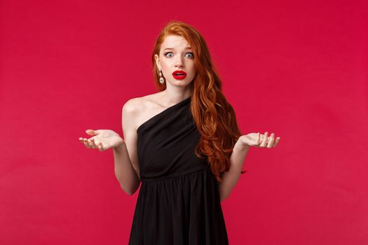Fashion, luxury and beauty concept. Portrait of shocked and freak-out redhead woman in elegant black dress telling police about what happened, raise hands sideways and look embarrassed.