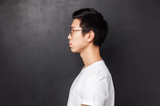 Profile portrait of serious-looking young adult asian man in white t-shirt and glasses, looking focused at left side of copy space, standing over black background no emotions, calm and relaxed.