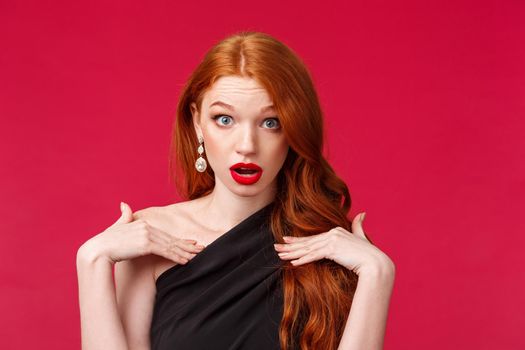 Close-up portrait of shocked gorgeous redhead woman look with disbelief and shook, questioned pointing at herself, being accused or blamed in something, stand in black dress red background.