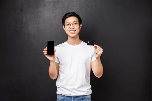 Bank, finance and payment concept. Portrait of simple asian guy in white t-shirt introducing new application for banking users, holding credit card and mobile phone, smiling camera black background.