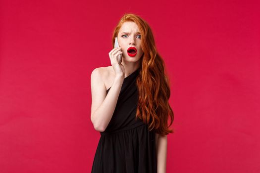 Portrait of disappointed and shocked gasping redhead girl in stylish black dress, hear from boyfriend he cant come, talking on phone and being displeased and let down, stand red background.