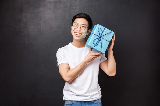 Celebration, holidays and lifestyle concept. Excited and curious happy young asian man celebrating birthday, shaking b-day gift box intrigued whats inside, smiling guessing, black background.