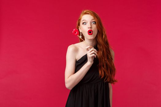 Portrait of elegant stylish redhead woman in black dress, folding lips in amusement and looking excited, holding lip cardboard, standing red background, having fun on girls party.