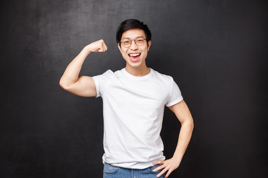 Proud and boastful handsome asian guy workout at gym, glex biceps to show girlfriend how strong he is, smiling pleased, do sports, exercise as having sportsman role-model, black background.