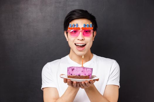 Celebration, holiday and birthday concept. Close-up portrait of excited and amused asian young man look amazed at candle on b-day cake, making wish, wear funny party glasses, black background.