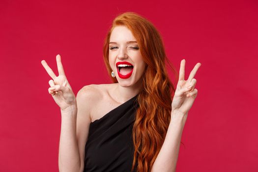 Close-up portrait of sassy and carefree redhead girl having fun, enjoying awesome party, show kawaii peace signs dancing joyfully, celebrating holiday in black dress and red lipstick.