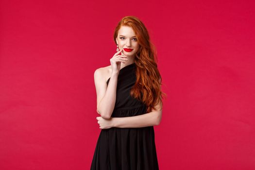 Elegance, fashion and woman concept. Portrait of seducative good-looking redhead woman in slim elegant black dress, red lipstick and evening makeup, look daring and sassy, red background.