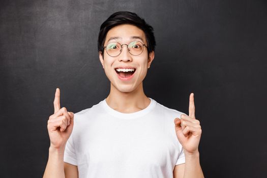 Close-up portrait of happy astonished and excited young asian guy seeing miracle something really awesome, pointing fingers up, smiling impressed and stare astounded at camera.