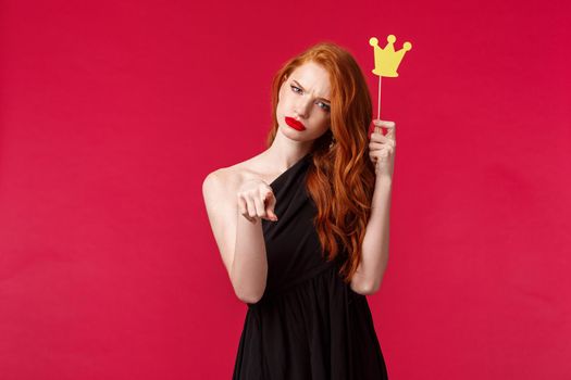 You mess with queen. Daring and sassy good-looking redhead woman in evening black dress, holding paper crown and pointing at camera, squinting serious or doubtful, red background.