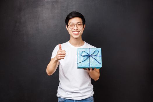 Celebration, holidays and lifestyle concept. Happy attractive asian man with present, holding birthday gift and smiling at camera with thumbs-up, guarantee you will like this thing.