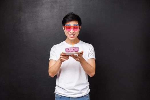 Birthday, celebration and party concept. Portrait of cheerful young asian male celebrating b-day blowing out candle on cake and smiling while making wish, thinking what he dreams about.