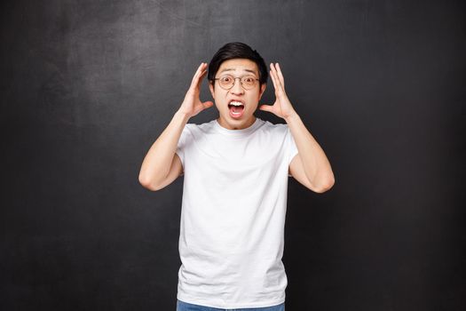 Alarmed and embarrassed young panicking asian guy, shaking hands near face as telling something shocking happened, retell outstanding story, staring camera, black background.