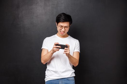 Technology, gadgets and people concept. Portrait of concentrated, focused asian guy trying kill mob in mobile game, passing arcade or street races, holding smartphone horizontally and grimacing.
