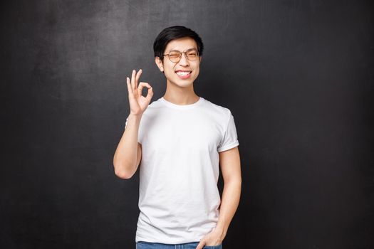 Carefree happy smiling young asian guy left satisfied after trying new product, visit company and use their good services, show okay sign and wink joyfully, pleased over black background.