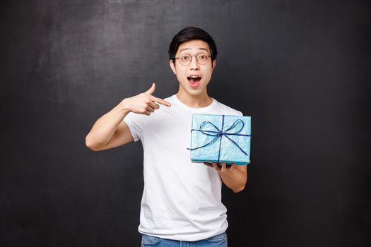 Celebration, holidays and lifestyle concept. Portrait of excited and curious cute asian guy asking whats inside gift box, celebrating b-day party holding present and pointing finger at it.