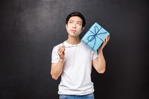 Celebration, holidays and lifestyle concept. Thoughtful and dreamy young handsome asian man celebrating birthday, thinking trying guess whats inside blue b-day box, stand black background.