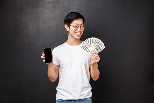 Technology, money and prizes concept. Pleased smiling happy rich asian guy in t-shirt, looking satisfied at dollars fan of cash and holding mobile phone, showing smartphone screen, black background.