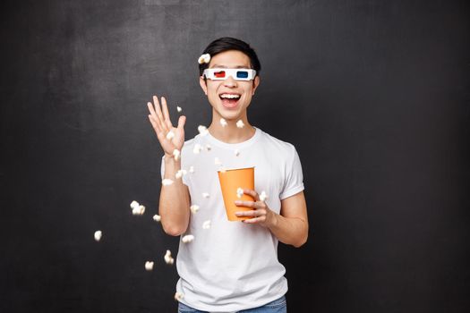 Leisure, movies and lifestyle concept. Portrait of amused and carefree funny asian guy in t-shirt throwing popcorn at cinema screen as watching movie 3d glasses, stand black background.