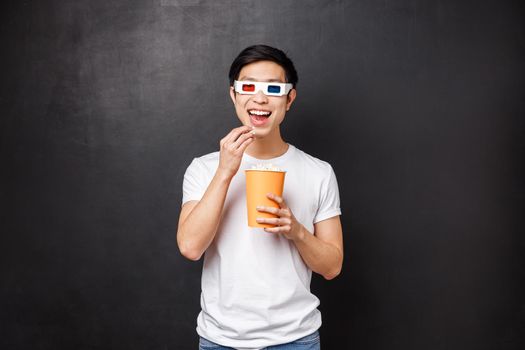 Leisure, movies and lifestyle concept. Amused and interested asian guy in 3d glasses smiling intrigued as watching awesome new movie, eating popcorn in cinema, black background.