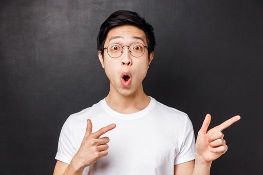 Close-up portrait of thrilled and amused asian young man in white t-shirt, open mouth amazed say wow, pointing fingers right at something impressive and stunning, white background.