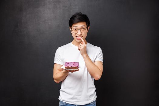 Potrait of shy and silly asian guy suggesting his piece of cake to girl he likes, asking try it as made dessert on his own, biting finger and look awkward at camera, asking your opinion.