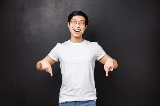 Happy pleased asian male customer bragging friends, showing his new product he boutght during discount holiday prices, pointing fingers down smiling satisfied and laughing at camera, black background.