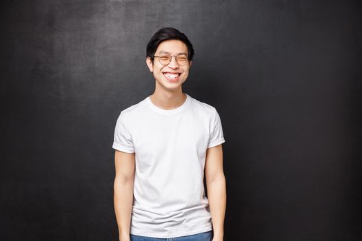 Portrait of cheerful asian guy with beaming white smile in t-shirt and glasses, looking camera laughing, express happy positive emotions, have carefree conversation, black background.