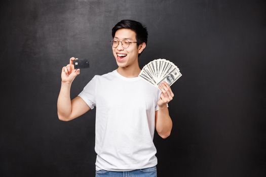Technology, money and prizes concept. Portrait of boastful smiling, happy asian guy become rich, lucky to win prize, holding dollars and credit card, making decison how invest money, black background.