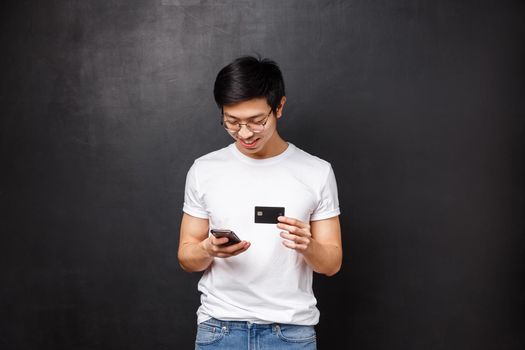 Bank, finance and payment concept. Young asian guy in t-shirt, holding credit card as insert billing info at shopping application, look at mobile phone display smiling, buying new clothes internet.