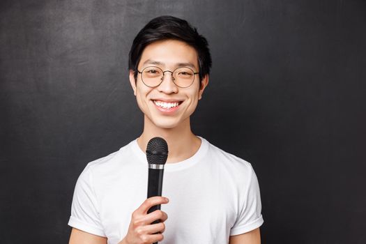 Leisure, people and music concept. Handsome and cute smiling asian man in white t-shirt, glasses, holding microphone, singing at karaoke party with friends, picking song on screen, black background.