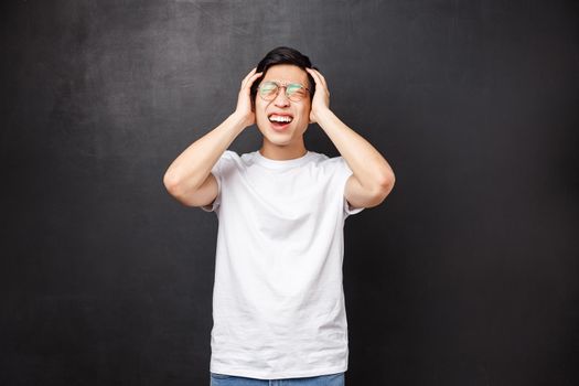 Depressed and tensed young asian guy fed up with studying, have lots on mind, grab head grimacing from pain feel distressed and upset, express anger and bothering feelings, black background.