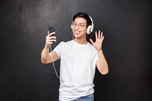 Technology, gadgets and people concept. Portrait of friendly smiling handsome asian man in t-shirt, wear headphones, waving hand say hello as talking on video-call using mobile phone contact friend.