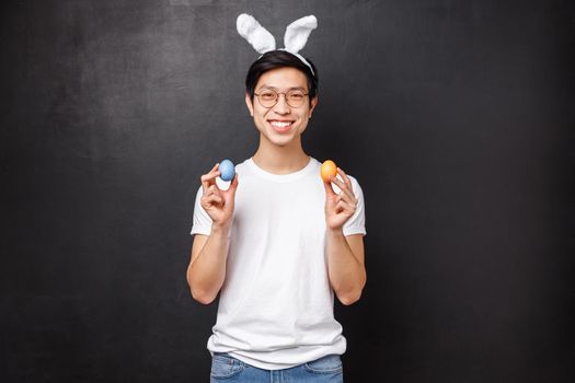 Holidays, party and Easter concept. Portrait of happy smiling friendly asian guy in rabbit ears celebrating orthodox Holy day, holding painted eggs and looking camera, stand black background.