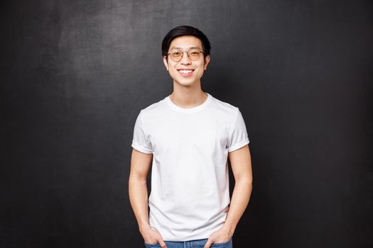 Portrait of young asian guy in glasses standing in white casual shirt over black background, friendly smiling, express happy enthusiastic emotion, hanging with mates after college.