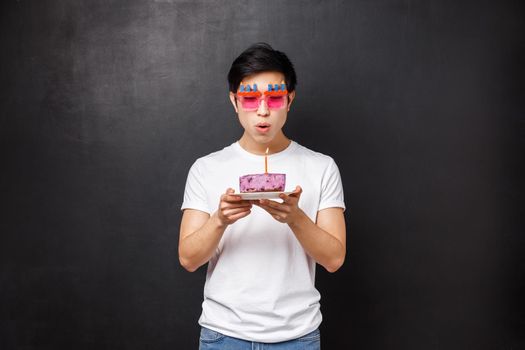 Birthday, celebration and party concept. Portrait of young asian guy grew up, wear funny sunglasses celebrating b-day blowing out candle on cake to make wish, standing black background.
