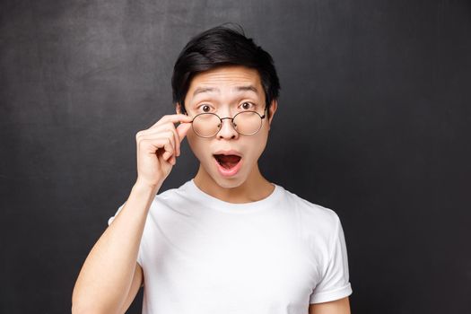 Close-up portrait of excited and wondered, intrigued asian man touch glasses, look excited and interested at camera, open mouth fascinated, listen to interesting suggestion, black background.