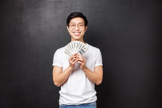 Portrait of lucky satisfied young rich asian male student holding lots of cash, fan of dollars smiling happy and pleased as winning prize, receive reward or his first paycheck, black background.