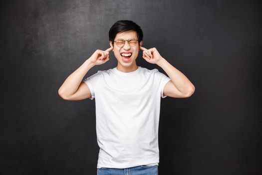 Lifestyle and people concept. Bothered and anguished young asian guy screaming from discomfort and distress as hearing loud noise, close ears with fingers shut eyes disturbed, stand black background.