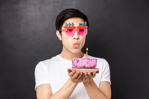 Celebration, holiday and birthday concept. Close-up portrait of dreamy cute asian man in funny party glasses, blowing out candle to make wish, dreaming on b-day, stand black background.