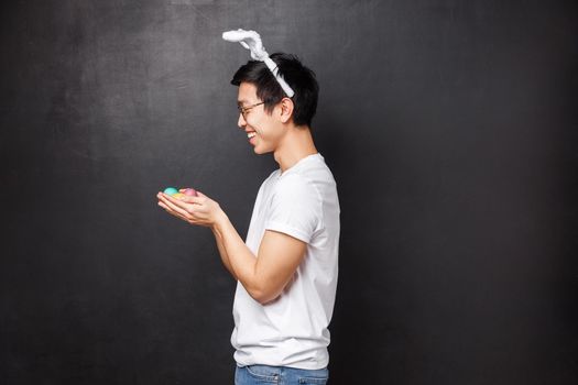 Holidays, party and Easter concept. Profile portrait of cheerful cute young asian man in rabbit ears and white t-shirt, standing over black background with painted eggs in hands.