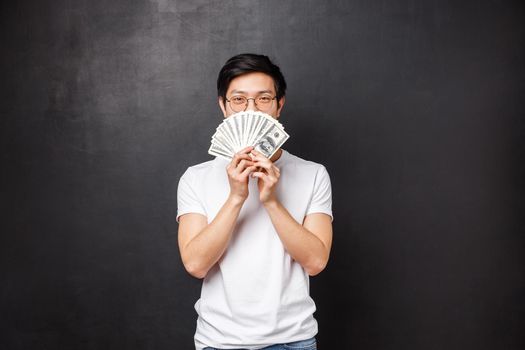 Portrait of sly happy and pleased asian young man winning big cash prize money, hiding face behind fan of dollars smiling with eyes, decide what to buy on it, standing black background.