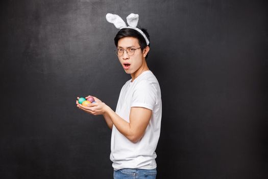 Holidays, party and Easter concept. Profile portrait of sassy funny and playful asian young man turn to camera in rabbit ears and holding painted eggs in hands, stand black background.