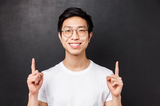 Close-up portrait of successful pleased young asian male entrepreneur, pointing fingers up and looking camera with satisfied beaming smile, talking about how to earn money, black background.