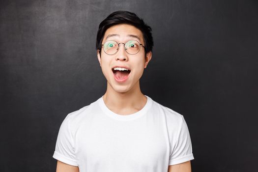 Close-up portrait of amused and wondered, enthusiastic asian man in white t-shirt and glasses, look astonished with impressed and excited face, open mouth wondered, stand black background.