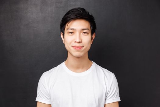 Close-up portrait of smiling friendly-looking young asian male model in white t-shirt, looking at camera and grinning, standing over black background, concept of people emotions and lifestyle.