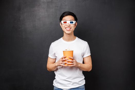 Leisure, movies and lifestyle concept. Handsome cheerful and excited asian man in 3d glasses, holding popcorn and smiling amused as watching favorite thriller or comedy, black background.