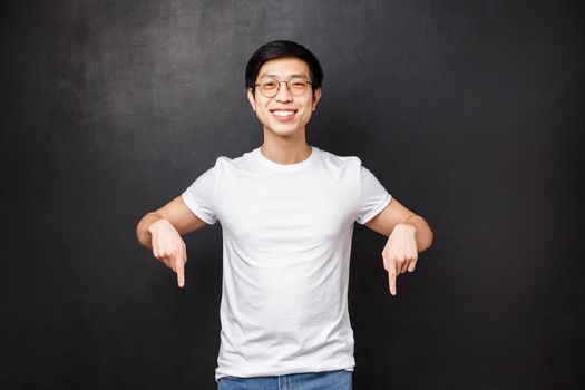 Studio portrait of happy handsome asian male student in white t-shirt, inviting new members join his team start career in business, pointing fingers down look here sign, smiling camera.